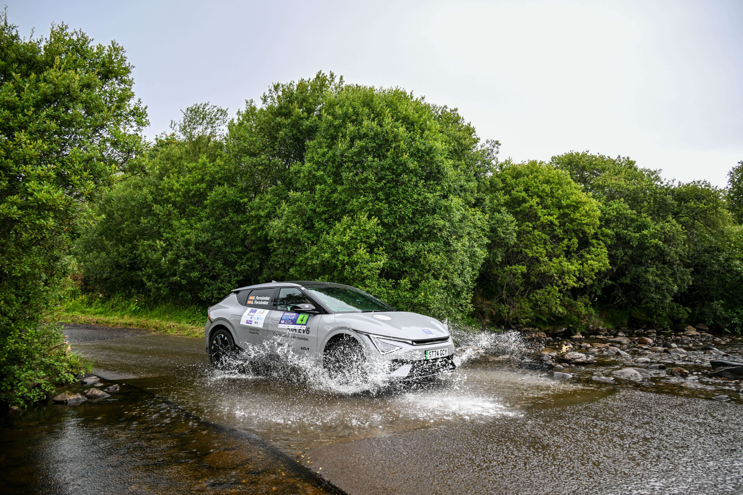 EV Driving through puddle on road on sunny day with lush green trees behind